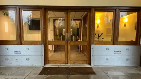 Julia Gregory/ BBC Brown external doors with glass panes. There are large metal panels in front of windows either side of the door