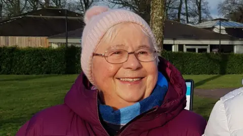 Gwynneth Exall wearing a blue and floral top, wearing glasses and with short grey hair, smiling at the camera, while sitting in a garden