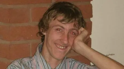 A man in a striped shirt with long blonde hair smiling at the camera, with his hand resting on his face