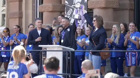 Stuart Howells/BBC Two people are holding up a trophy, that is silver with purple ribbon, with one hand each. Ipswich Town Women members are standing around them wearing blue T-shirts and clapping.