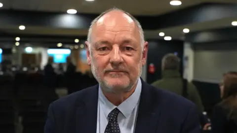 John Fairhall/BBC Simon Pepper with short grey hair. He is wearing a dark blue suit, a light blue shirt and a dark blue patterned tie. He is looking at the camera with other people facing away in the background.