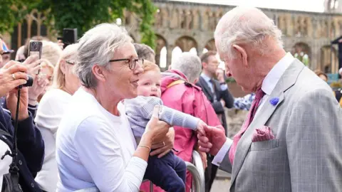 PA Media King Charles shakes the hands of a little child with red hair being held up by a woman with grey hair and glasses in a lilac jumper
