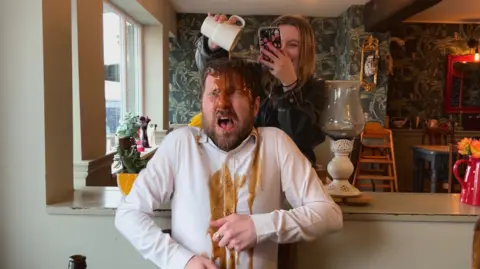 A man in a white shirt is sitting in a pub. A woman holds a camera phone and pours gravy over his head from a white cup. Lamps, plants, flowers and leafy wallpaper decorates the pub.