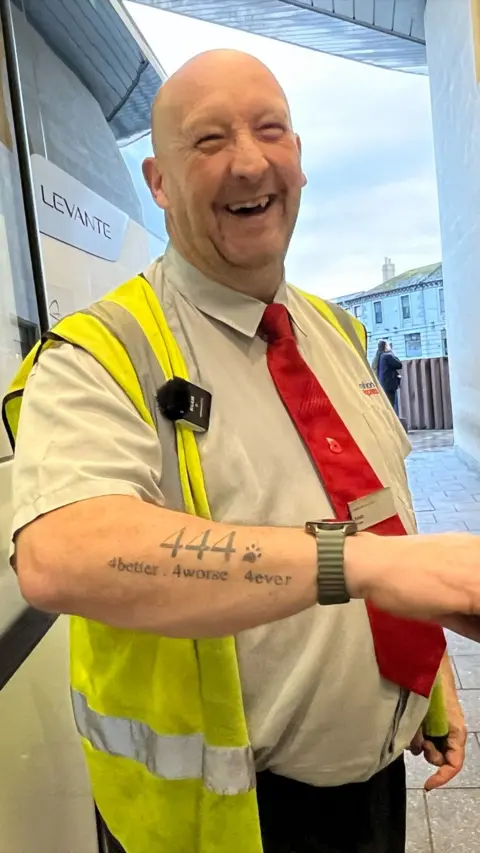 Man smiling into the camera with a red tie and showing his tattoo to the camera that is the numbers '444'.