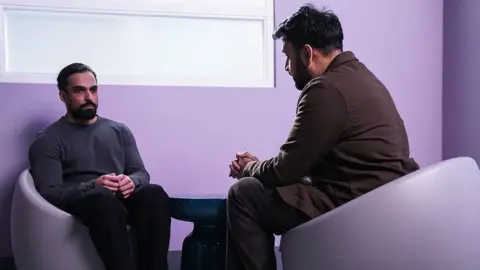 BBC/Jack Barnes/Kieron McCarron A still from EastEnders, showing Ravi speaking to a mental health professional, sitting on purple seats in a purple room.