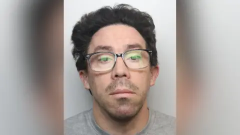 Shaun Smith has medium length slicked-back black hair, dark stubble and glasses.