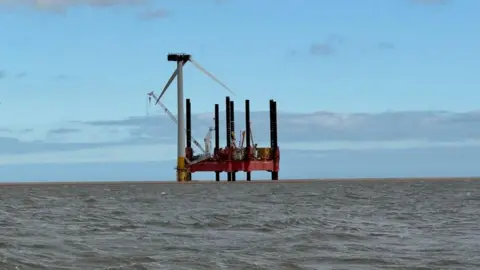 Andrew Turner/BBC A wide angle image of the burned turbine showing one blade and the housing removed on one turbine within the offshore wind farm. The uppermost blade has been removed, with two in the downward position. A jack-up barge is visible featuring a crane jib and the deck and struts of the barge. The sky behind the semi-silhouetted image is blue and the sea is brown-ish.