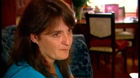 BBC A woman with dark long hair sits on a blue sofa with a table and chairs behind her. She has brown eyes and is wearing a light blue top. 