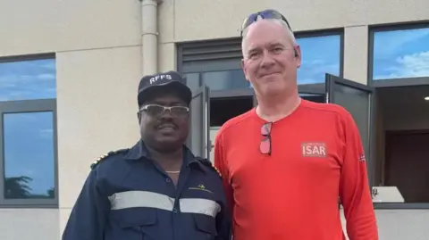 LFB A man in red stands with a man and black and grey. They both smile at the camera proudly.