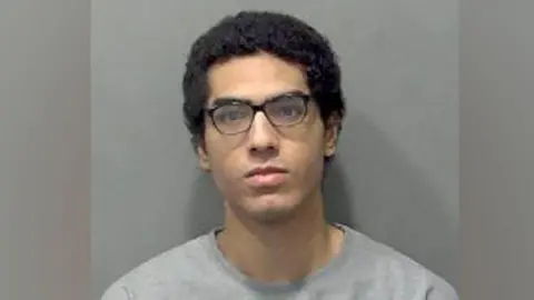 Nicholas Prosper, who has dark hair and dark glasses, staring into the camera for a police custody image. He is wearing a grey sweatshirt.