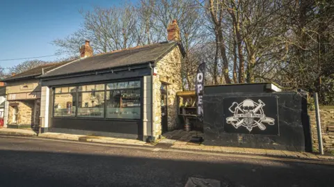 The Bearded Brewery The Bearded Brewery shop in St Agnes on a sunny day. It sits on the roadside and is a small single storey building with a large glass window front. Its logo is painted on a wall nearby and it is a black and white skull and crossbones.