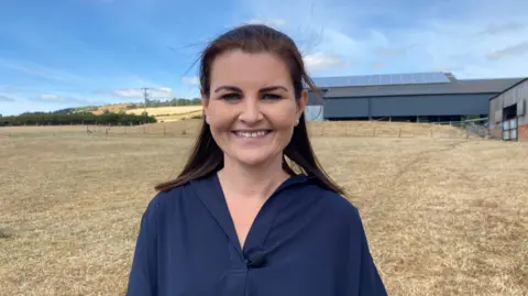 Ellen Knight/BBC Photo of Jones looking into the camera and smiling. She is wearing a dark blue top, and is stood in front of a farm barn. The field she is stood on is grassy, but has yellowed in the sun. The sky is blue and there is strong sunshine. 