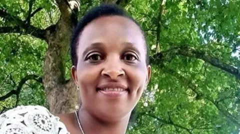 Irene Mbugua has short black hair. She is smiling at the camera. We can see there is a tree behind her. The photo has been taken from below her so she is looking down. 
