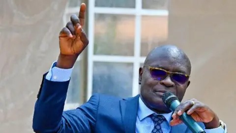 Simon Byabakama in glasses, wearing a blue suit and a dotted tie and speaking into a mic 