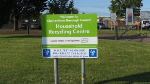 A green sign on two metal poles on a large grass verge that reads "Welcome to Cheltenham Borough Council, Household Recycling Centre". The recycling centre and trees can be seen in the background on what is a sunny day.