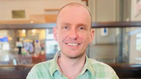 Ben Howard, a man with a buzz cut and stubble, is pictured smiling at the camera. He is wearing a light green, checkered shirt with a collar. He appears to be sat in a pub. 