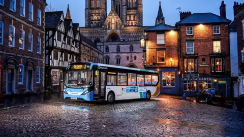 A Stagecoach bus with blue, white, green and orange markings on cobbled pavements in front of Lincoln Cathedral. The bus is parked at an angle. It is evening, so it is getting dark. There are buildings surrounding the bus with lights on. 