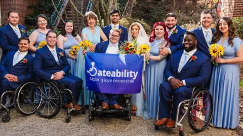 A posed shot of a group of people at a wedding. The groom is in a wheelchair and is holding a banner saying 'Dateability - making love accessible'. The bride is standing next to him holding a large bunch of sunflowers. There are several bridesmaids in long blue flowing dresses, and several male ushers, three of whom are also in wheelchairs