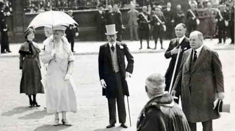 Merseytravel King George V and Queen Mary being welcomed by notable figures at the tunnel entrance. He is holding a cane and wearing a top hat and tails. She has a white parasol and matching ornate dress. 