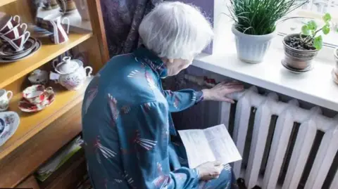 An elderly woman with white hair huddled up to a radiator holding a bill 