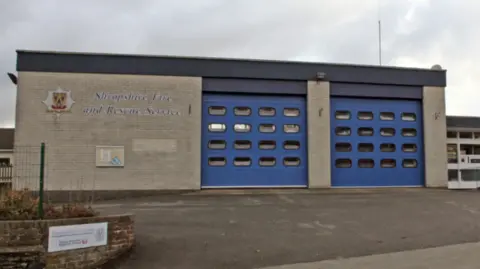A fire station set back from the road with two large blue doors. The words Shropshire Fire and Rescue Service are in large letters on the building to the left of the doors on the photo.