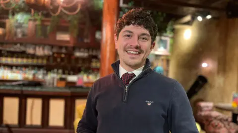 A mid shot of Xavier Leguitte, who is wearing a black jumper and red tie, and is smiling