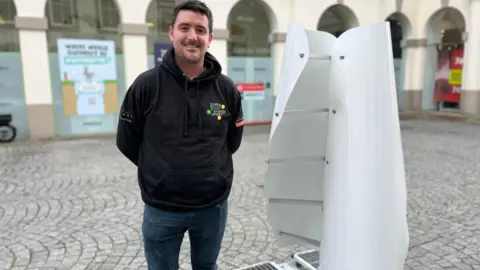 A man stands next to a white wind turbine, which is made of two curved, vertical sails on a flat base. The turbine is slightly shorter than the man.