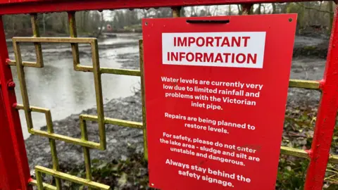 A red sign is affixed to a red and gold gate saying Important Information - Water levels are currently very low due to limited rainfall and problems with the Victorian inlet pipe. Repairs are being planned to restore levels. For safety, please do not enter the lake area - sludge and silt are unstable and dangerous. Always stay behind the safety signage. 