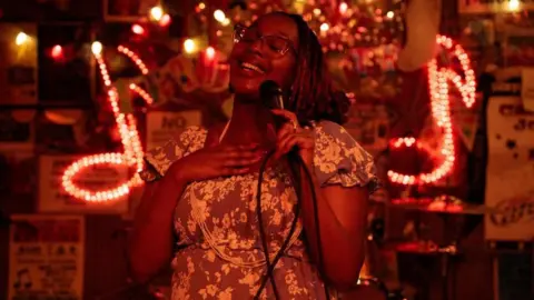 Edna Nicole Luckett sings at a microphone, her hand on her heart, with light-up signs showing musical notes behind her