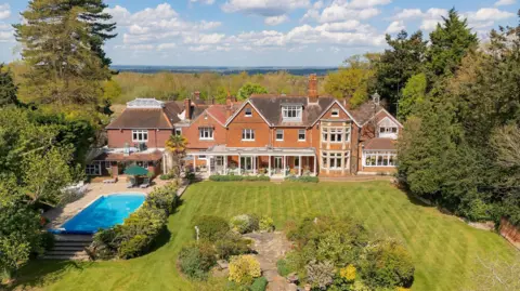 Savills A huge house can be see from a drone. It has a well kept green lawn and is surrounded by huge trees. There is also a swimming pool on the left hand side. The middle of the huge house has columns to hold up a roof creating an outdoor seating area under cover.