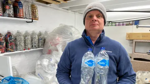 Richard Lord in a grey hat and a blue jumper holds two 1 litre plastic water bottles