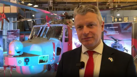 Luke Pollard is standing in front of a helicopter at Leonardos in Yeovil, wearing a black suit and red tie. He is smiling.