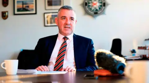 PFNI A grey haired man in a navy suit, white shirt and navy and red tie, sat at a wooden table with a white coffee mug and microphone on its surface. There are out of focus framed pictures on the wall and a PSNI green crest.
