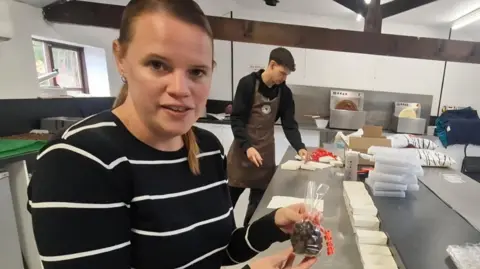 BBC/LIZ SAUL A woman in a stripey navy and white jumper, with long light brown hair, tied in a pony tail. She is standing in the kitchen of the chocolate factory, holding a clear bag of chocolate nuts. In the back ground, a young man is folding gift boxes from cardboard templates.