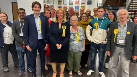 A group of people stand together, all wearing yellow Liberal Democrat rosette pins, in the centre is Liz Jarvis