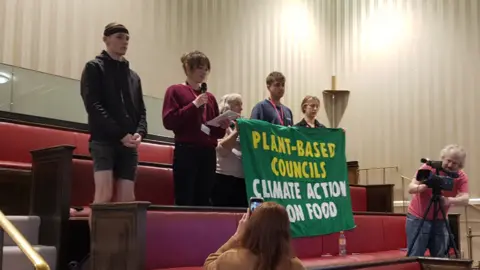 Image shows five vegan campaigners standing in the stalls at a council meeting, with three campaigners holding a green banner that reads 'PLANT-BASED COUNCILS CLIMATE ACTION ON FOOD' 