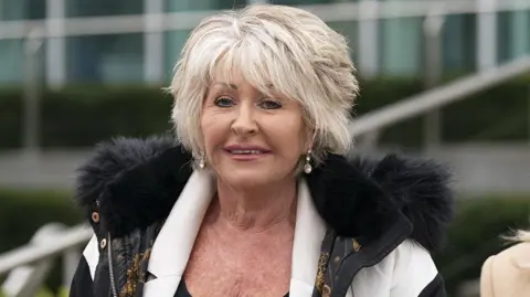 PA Media Former detective Maggie Oliver, who has short blonde hair, wears dangly earrings and a cream jacket with black furry hood. She is standing outside a large building.