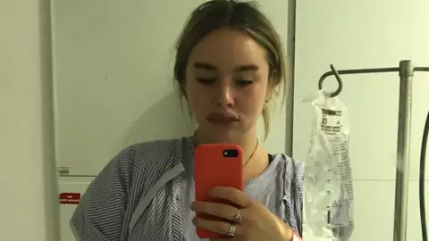 Chloe Dickson Chloe Dickson looks down at her phone while taking a selfie in a mirror. She is attached to a drip and is wearing a blue and white stripped hospital gown. The right side of her lower face is swollen following an anaphylactic shock. She stands against a plain white hospital wall with her drip and stand positioned to the left of her.