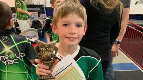 Tim Clarke Fin Clarke wearing a black and green bowling shirt and holding a small bronze coloured trophy.