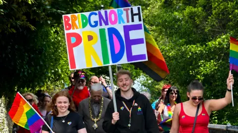 Photo of the 2025 Pride parade in Bridgnorth. At the front is Rees, who is wearing a black jumper with a rainbow heart on it. He is carrying a large sign, which reads 'Bridgnorth Pride" in rainbow letters. People in the parade are all waving rainbow flags and smiling. 