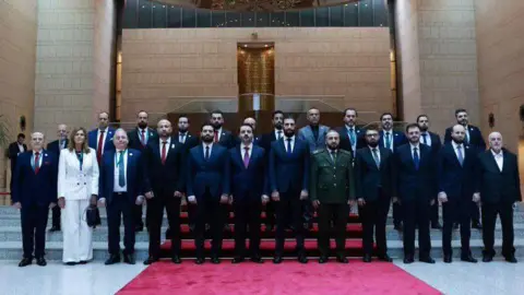 Syrian Presidency Members of the Syrian transitional government pictured standing in two lines, including President Ahmed al-Sharaa and Hind Kabawat. Kabawat is the only woman, and her white trouser suit stands out against the dark suits of her male colleagues.