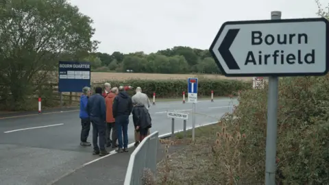 DJ McLaren/BBC A group of people, including inspectors, look out over Bourn Airfield, with a sign in the foreground denoting the location. 