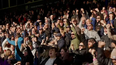 A packed scene of fans standing up, with arms raised, in the stadium.