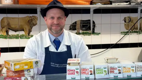 Butcher Dafydd Davies wears an apron and butchers coat and hat and stands next to his meat counter and a tiled wall with a pig, cow and sheep on the wall. 