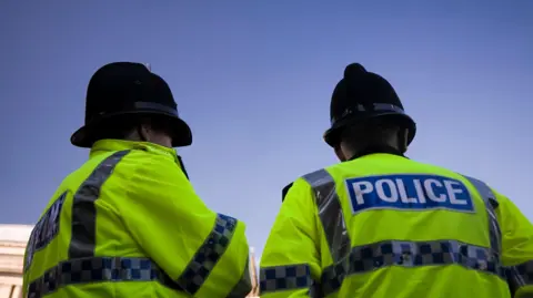 Two police officers with their backs in view, wearing police helmets and yellow high vis jackets with POLICE written across the back.