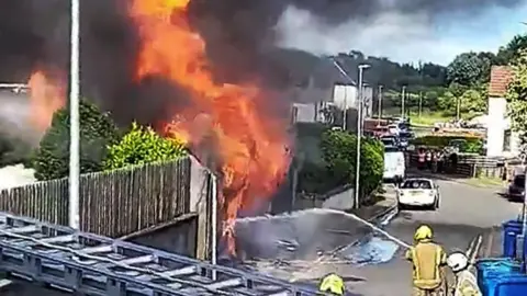Fife Jammer Locations A large orange fire on a street being extinguished by fire fighters with a hose with water dousing the flames.