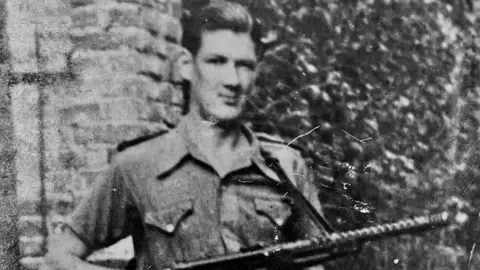Family Handout A black and white photograph shows a young man with slicked back dark hair looking at the camera. He is wearing a short-sleeved military shirt and carrying a rifle.