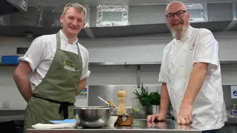 Two chefs stood in the center of a kitchen either side of a workspace. The man on the left, Harrison, is stood wearing a green apron with his hands behind his back. The man on the right, Simon, is wearing his chefs whites and is leaning on the workbench. 