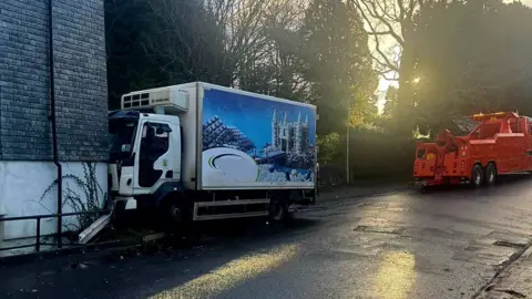 Buckfastleigh Fire Station The picture shows a roadside scene with two vehicles. On the left, there is a white refrigerated lorry, its cab is pressed against the wall of a brick building. Part of a metal railing appears bent or damaged near the impact point. The lorry has a large graphic on its side featuring an icy landscape with a castle.