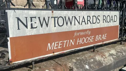 Street sign for Newtownards Road - 'formerly Meetin Hoose Brae' is written underneath.